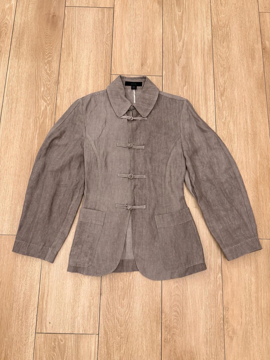 Light brown jacket with a collar, long sleeves, front pockets and knot fastenings, displayed on a wooden floor.