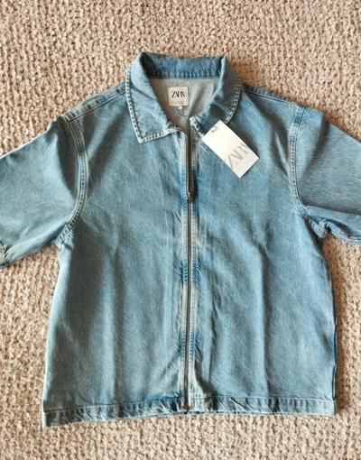 Light blue denim jacket with a front zip and a visible "ZARA" label on the collar, lying on a beige rug.