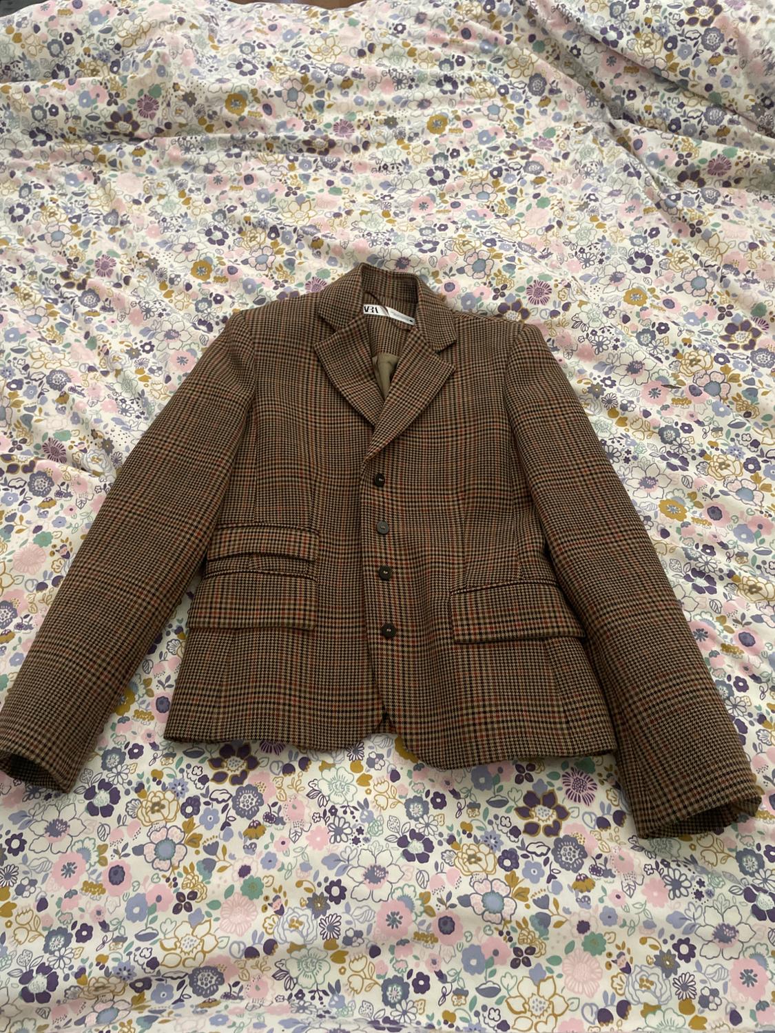 Brown checked jacket on a colourful floral bedspread. Visible label with text "ZARA".