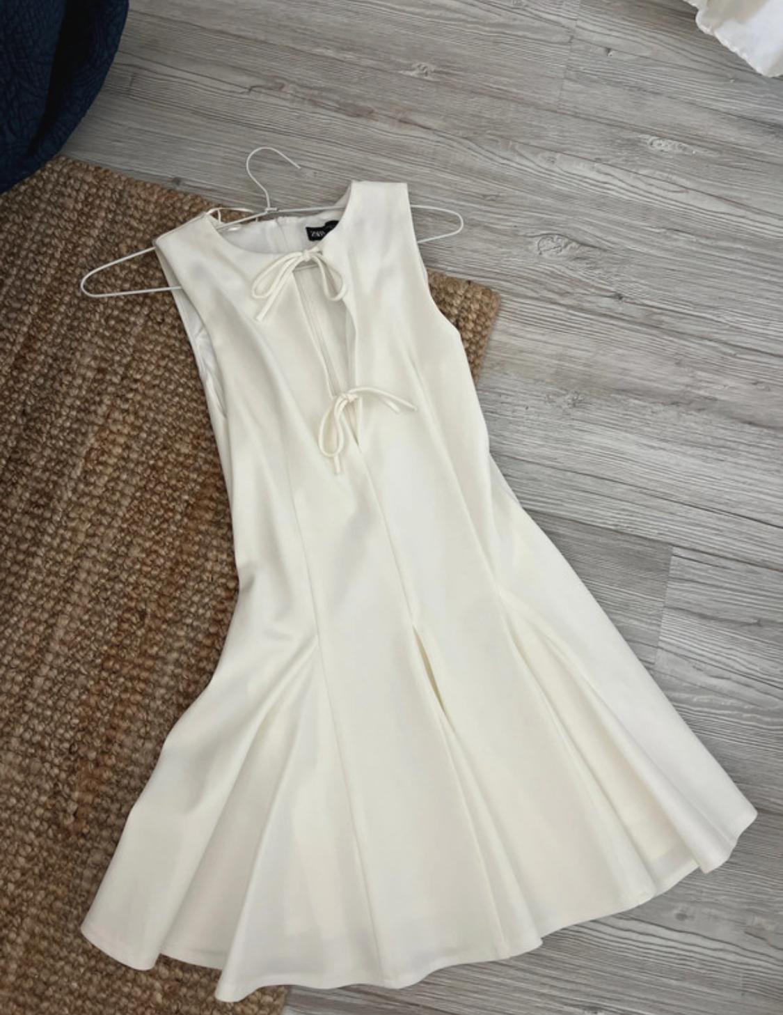 Short white sleeveless dress with front ties and ZARA label, on a hanger against a wooden floor and woven rug. Relaxed style.