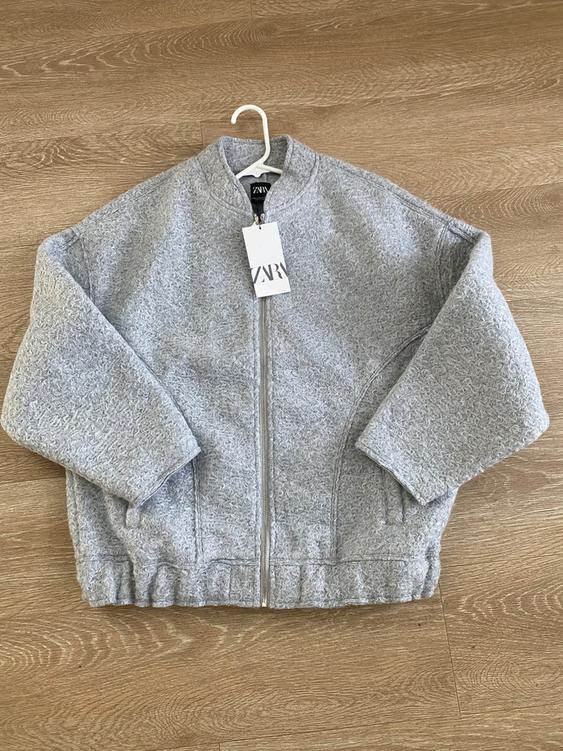 Gray jacket with zipper and round neck, hanging on a hanger over a wooden floor. Visible tag with text "ZARA".