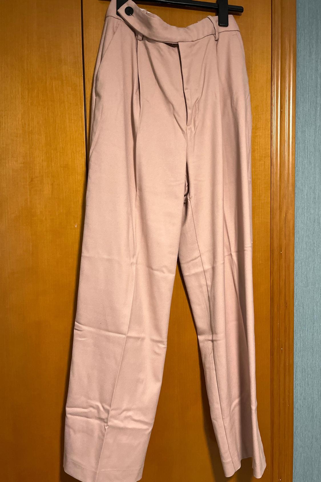 Front view of light pink trousers with pleats and a black button at the waist.