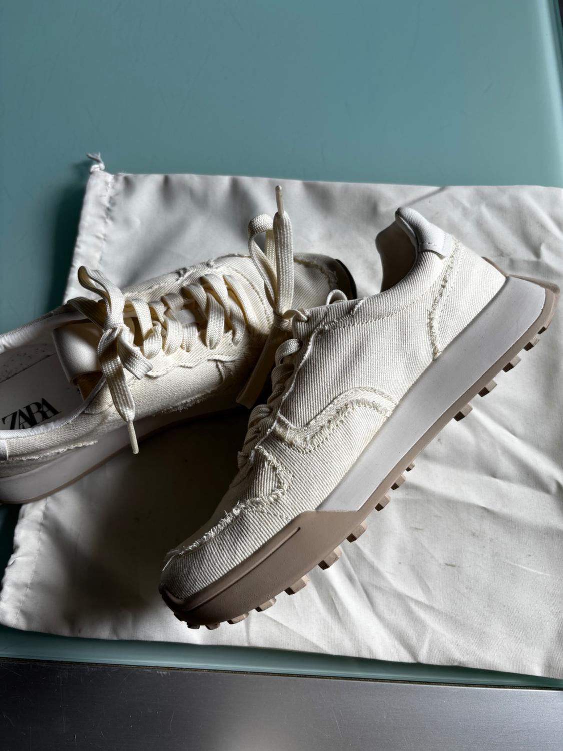 White trainers with a brown sole on a white fabric bag, placed on a blue surface. Visible text: "ZARA".