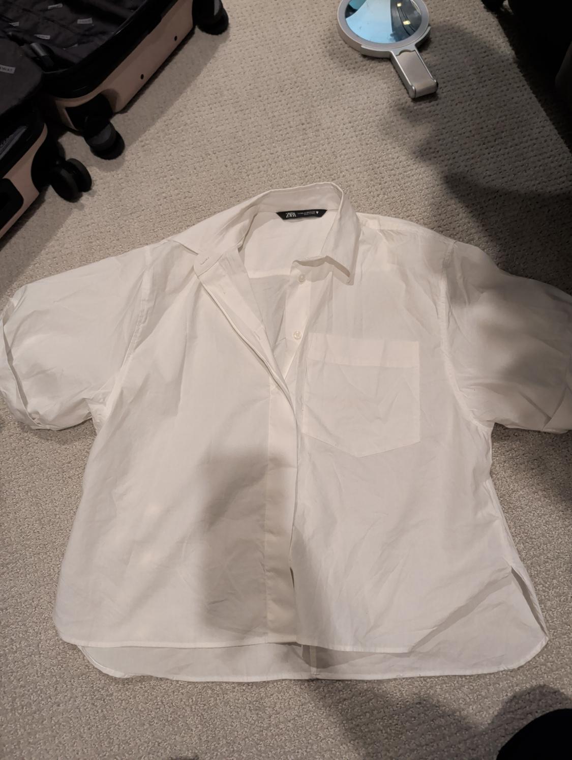 White short-sleeved shirt on a beige rug, with suitcases and a hand mirror around it. Visible label on the collar.