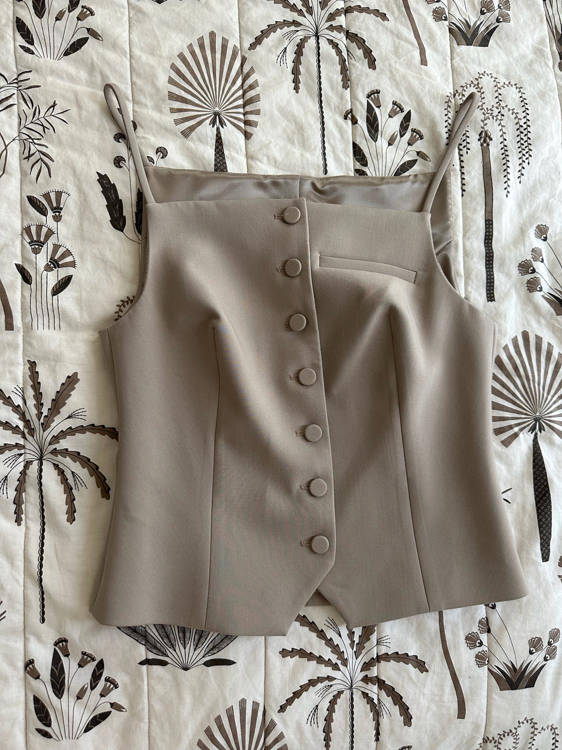 Beige camisole top with thin straps and front buttons, placed on a light-coloured bedspread with a brown palm-tree print.