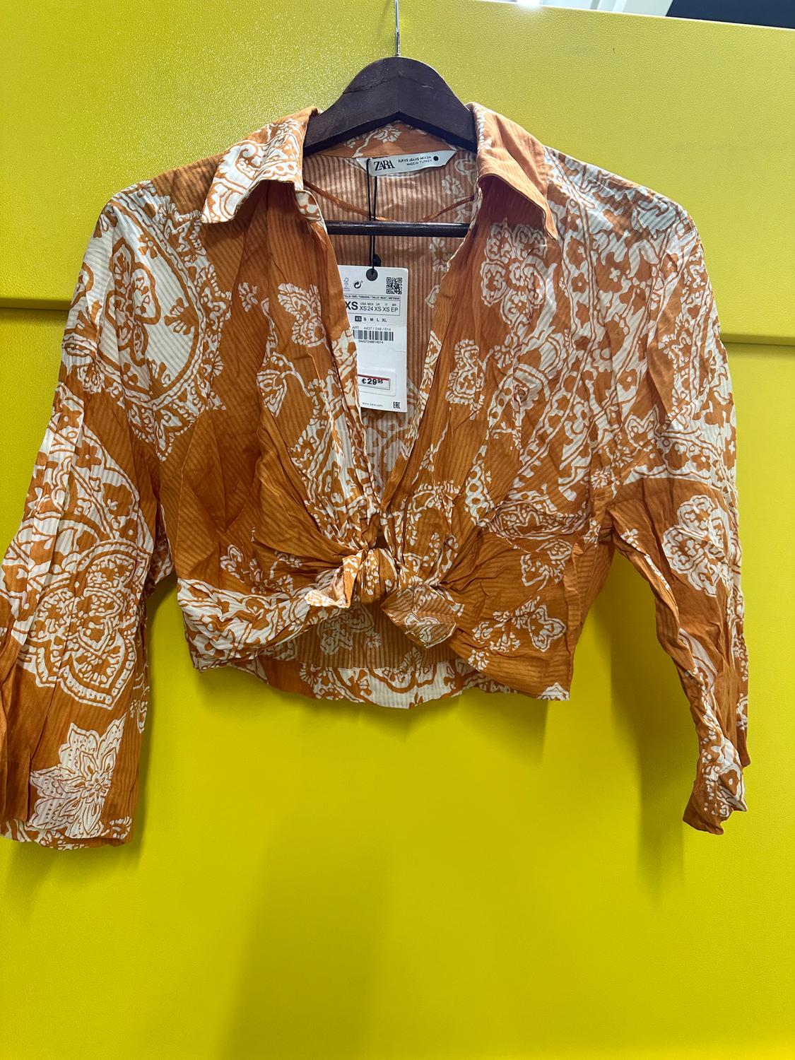 Brown blouse with white print and front knot, hanging on a hanger against a yellow background. Visible tag with price and size XS.