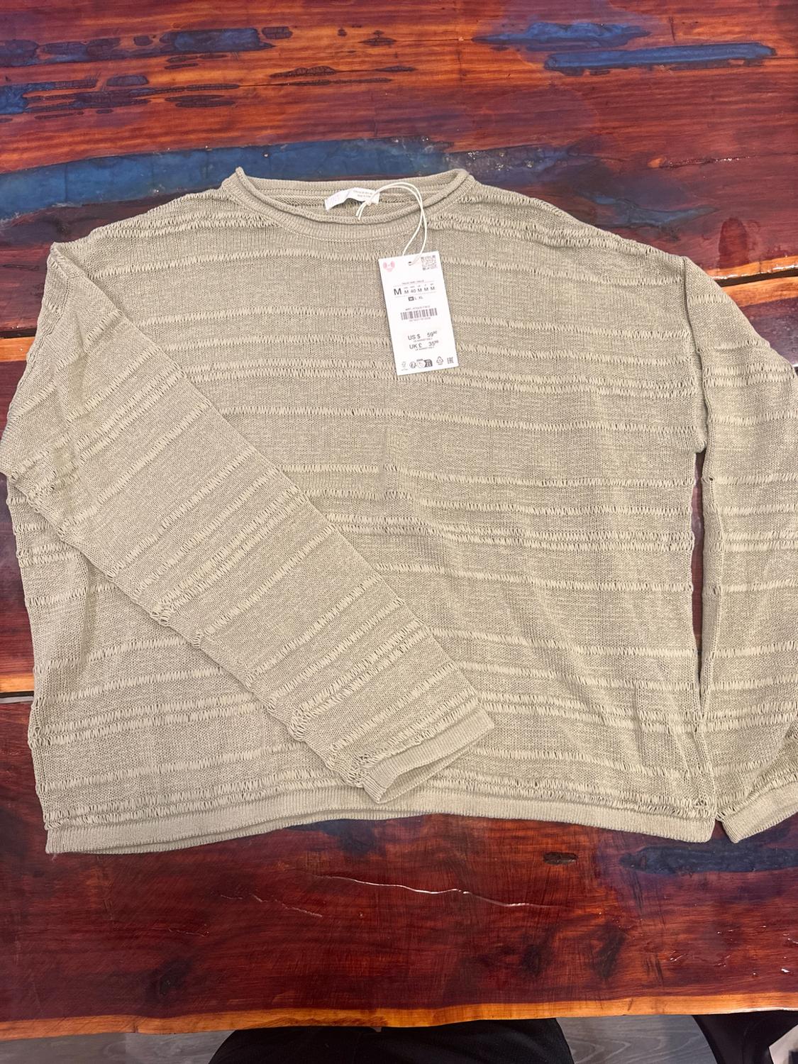 Beige knitted jumper with horizontal stripes and a label, on a rustic wooden table with blue and brown details.