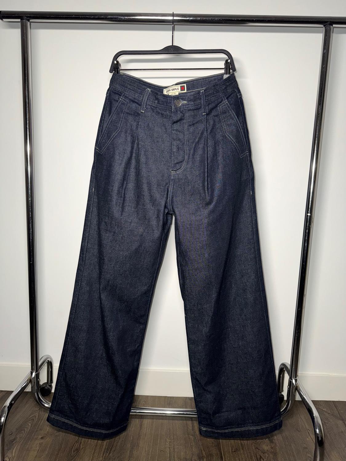 Dark blue denim trousers hanging on a metal clothes rack against a white background. Label visible at the waist.