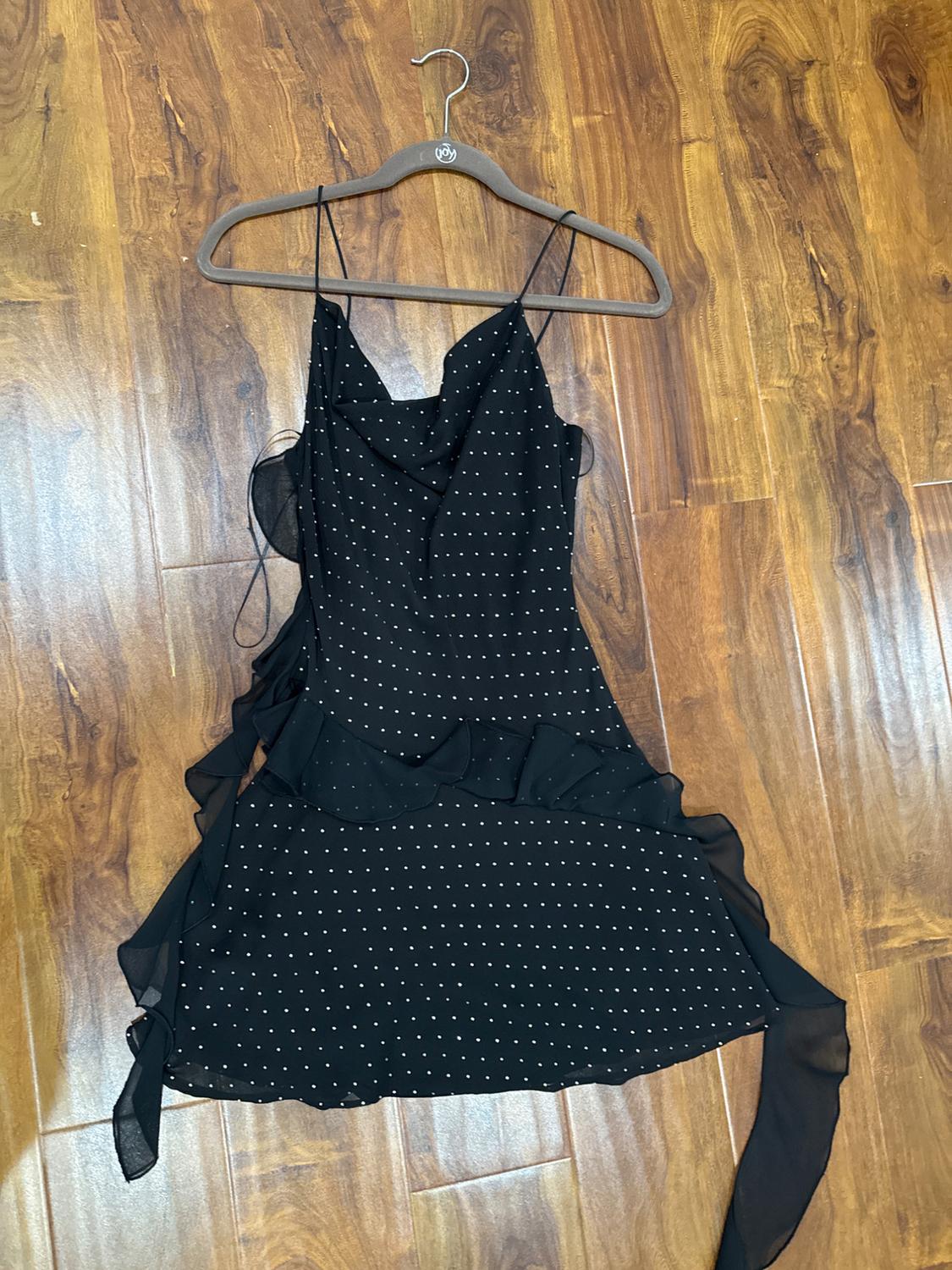 Black dress with spaghetti straps and frills, white polka dot print, hanging on a hanger over a wooden floor.
