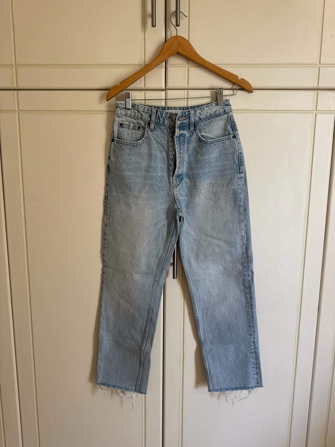 Light blue straight-leg jeans with a frayed hem, hanging from a wooden hanger in front of the doors of a white wardrobe.