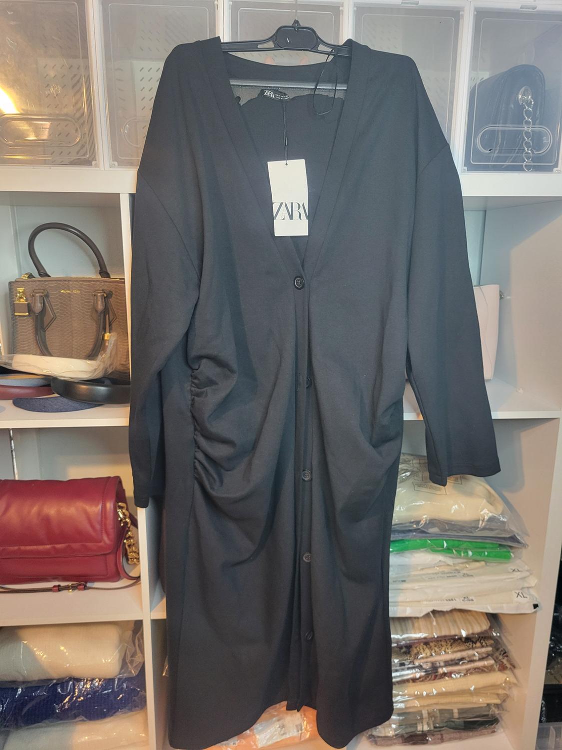 Long black coat with buttons hanging on a hanger, visible tag with text "ZARA", shelves with handbags and clothes in the background.