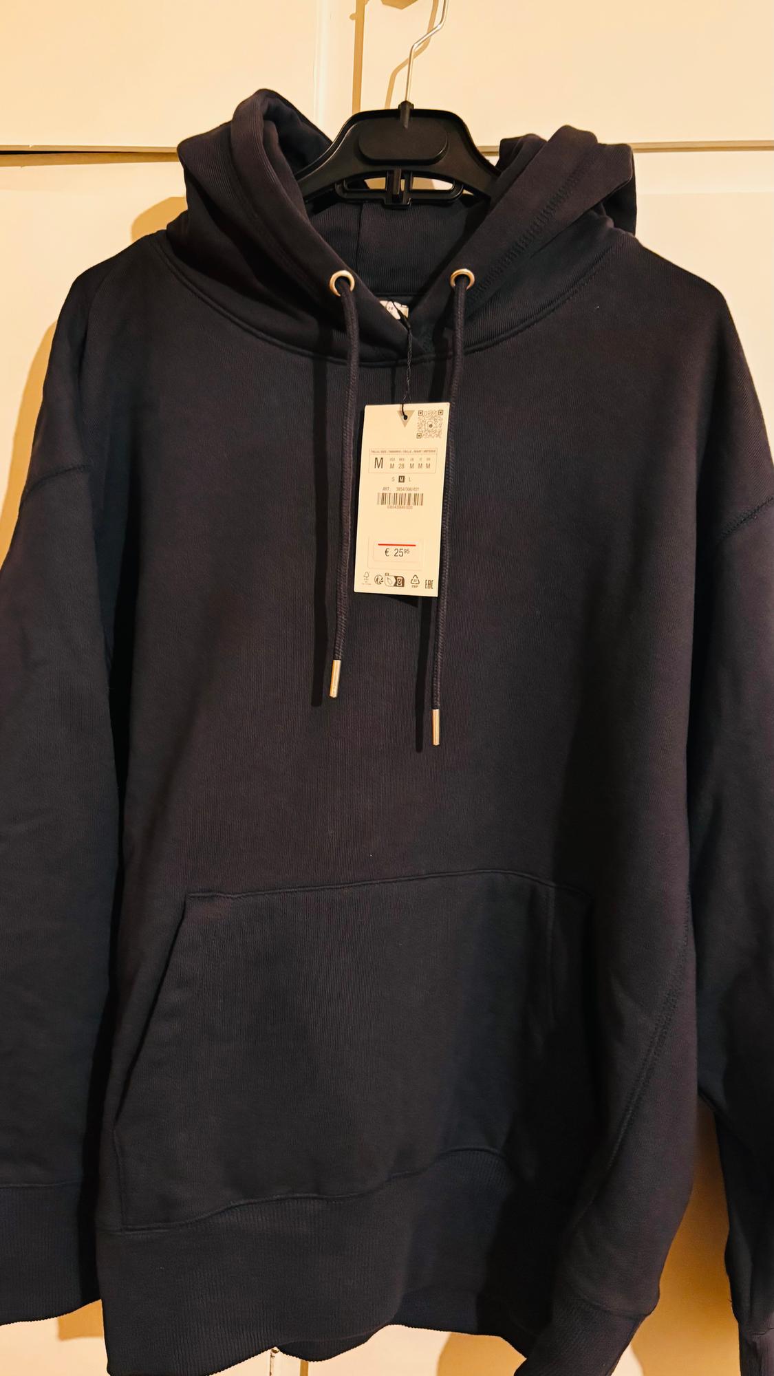 Black hoodie hanging on a hanger, visible tag with price of €25. White background.