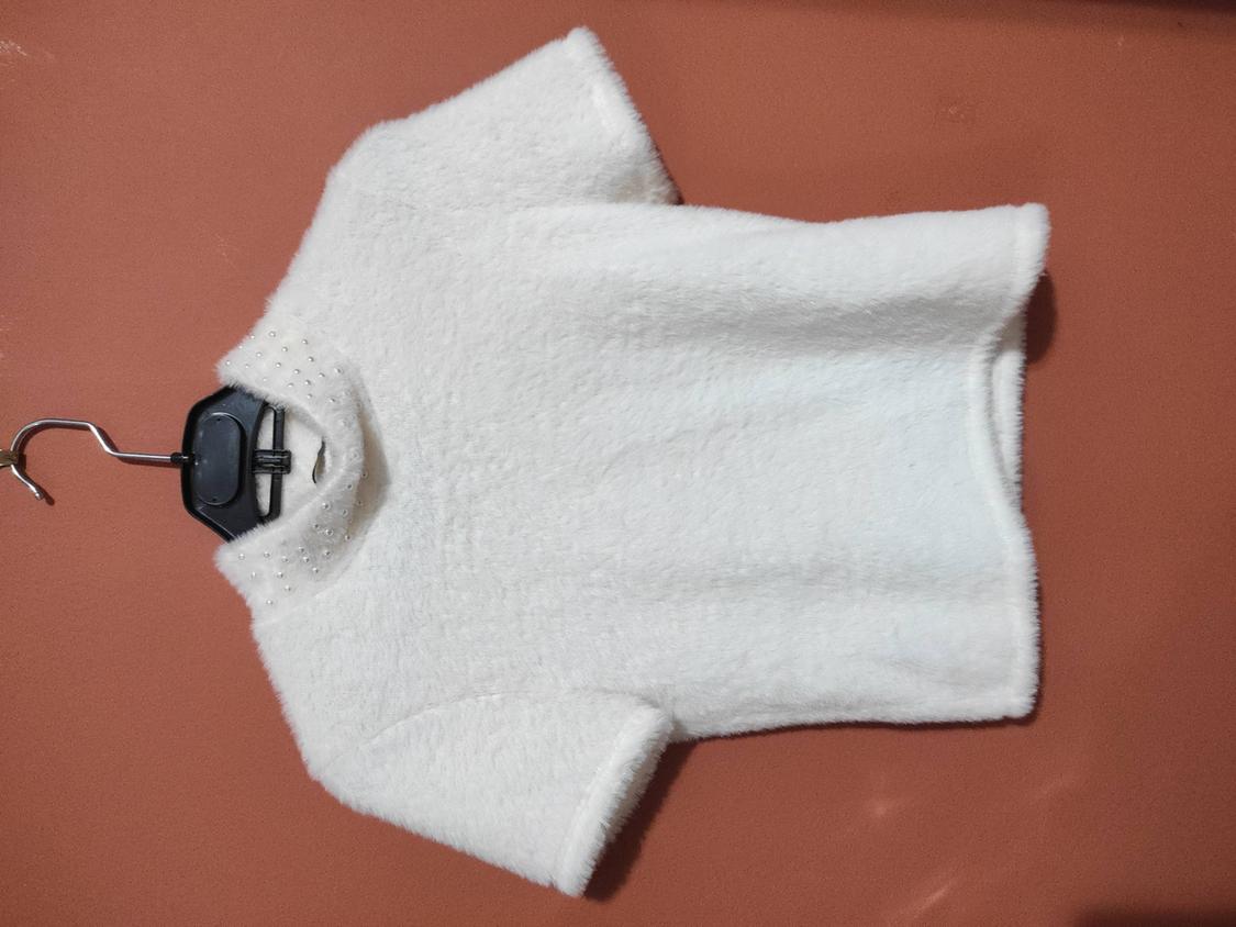White, short-sleeved t-shirt with a soft texture and a collar decorated with small pearls, hanging on a black hanger against a brown background.