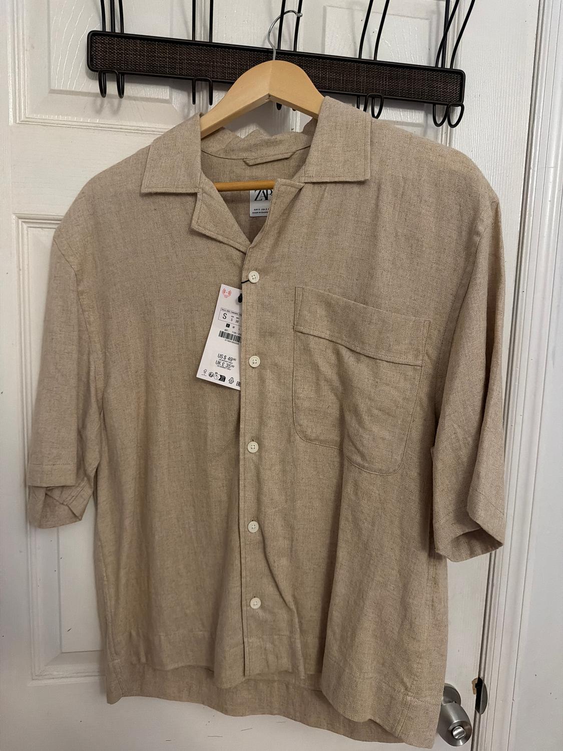 Beige short-sleeved shirt hanging on a wooden hanger, with a visible label. White door background with black coat rack.
