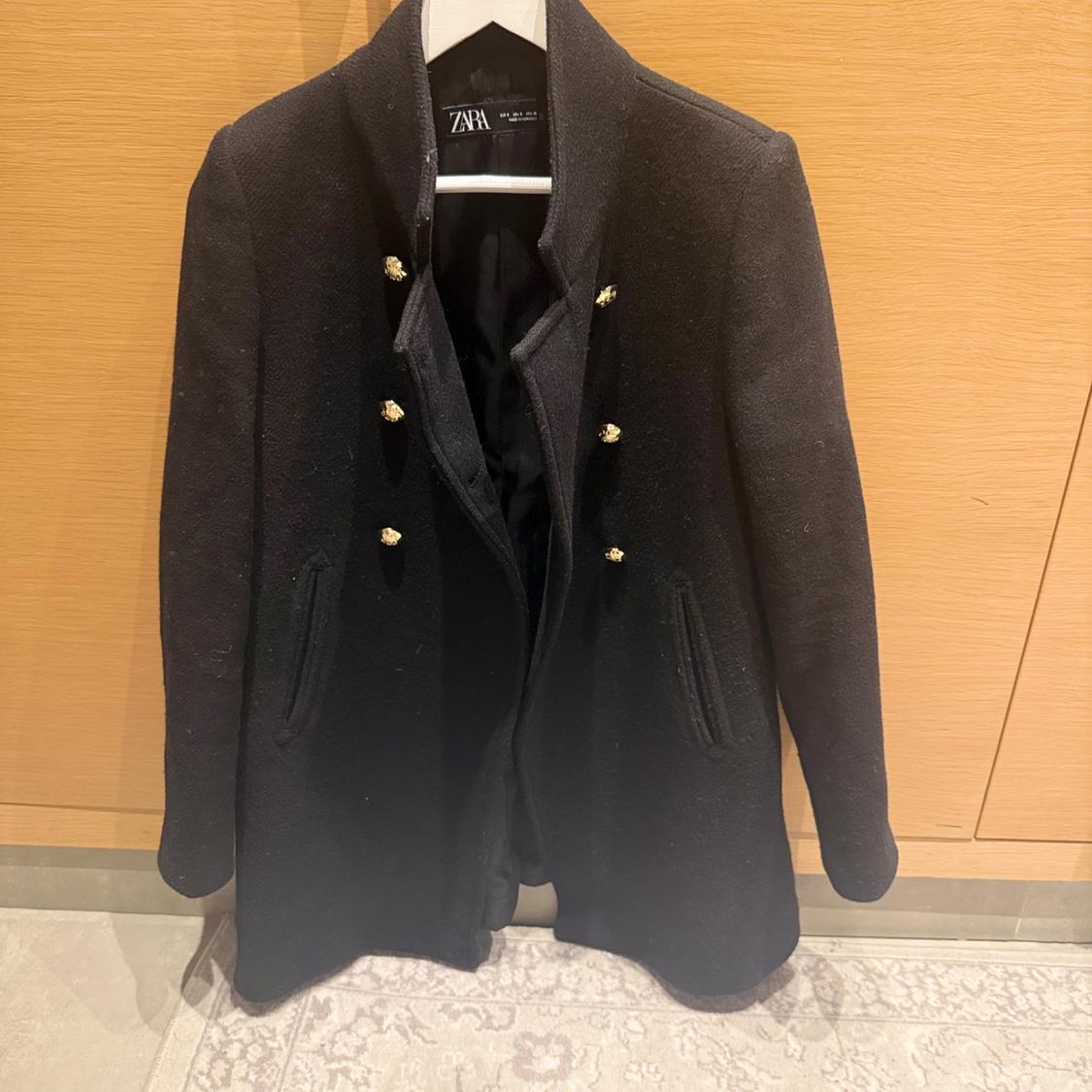 Black coat with gold buttons hanging on a wooden hanger, against a light wood background and beige carpet.
