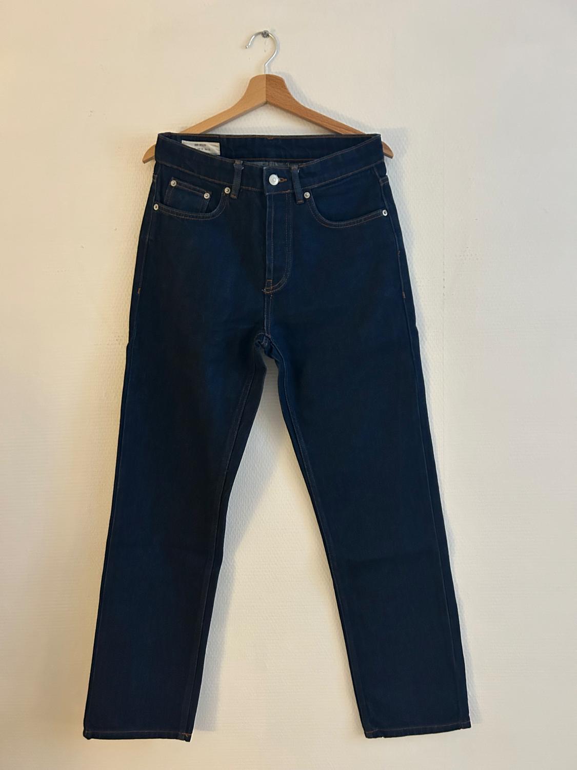Dark blue jeans hanging on a wooden hanger against a white wall. Classic five-pocket design with visible stitching.