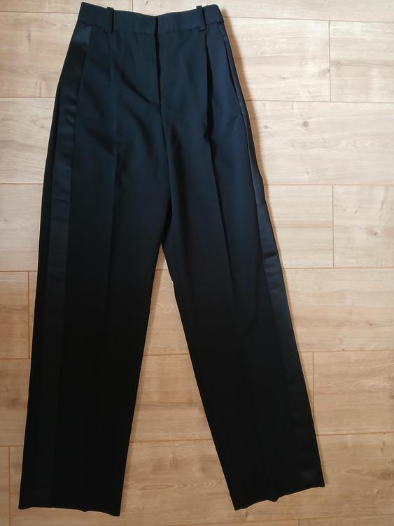 Black dress trousers with pleats, waistband with belt loops and side stripes, on a light wooden floor.