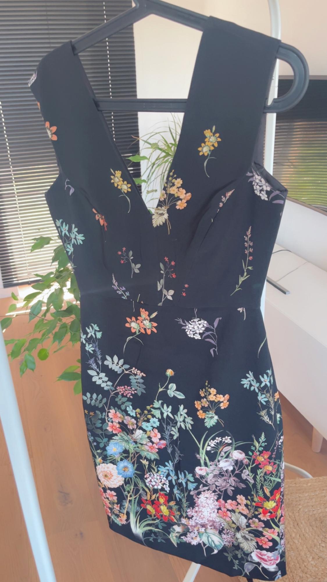 Black dress with a colourful floral print hanging on a hanger. Background with black blinds and a green plant.