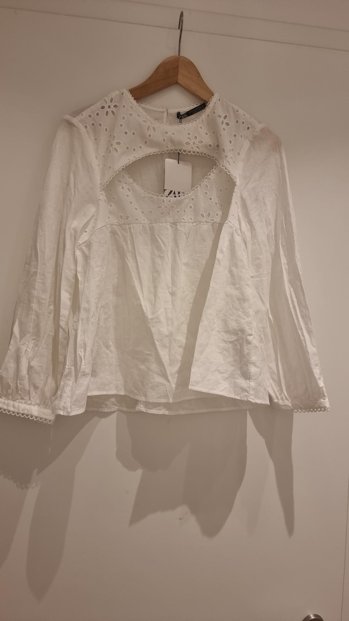 Long-sleeved white blouse with floral embroidery and a keyhole neckline, hanging on a wooden hanger against a white background.
