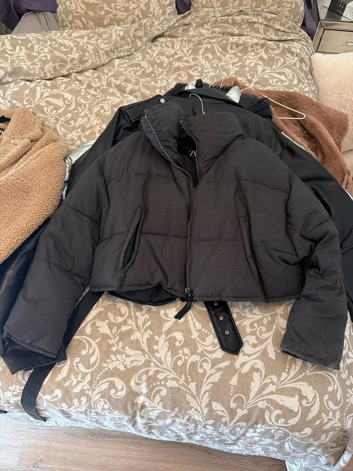 Black quilted jacket with zip and ZARA label on a bed with a patterned bedspread. Other black and brown garments are underneath.