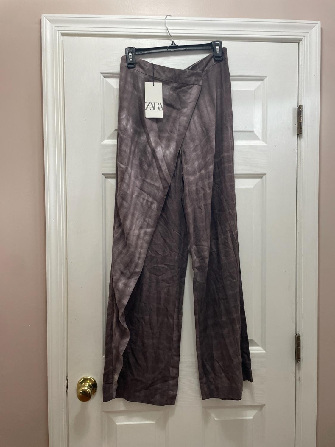 Flowy brown pants with a bleached effect and a crossover panel, hanging on a white door. Visible label with the text ZARA.