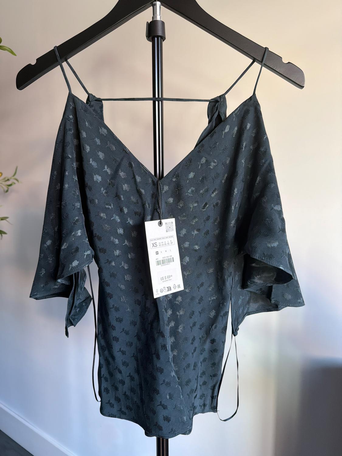Blue blouse with thin straps and subtle print, hanging on a black hanger. Visible tag with size XS and price details.