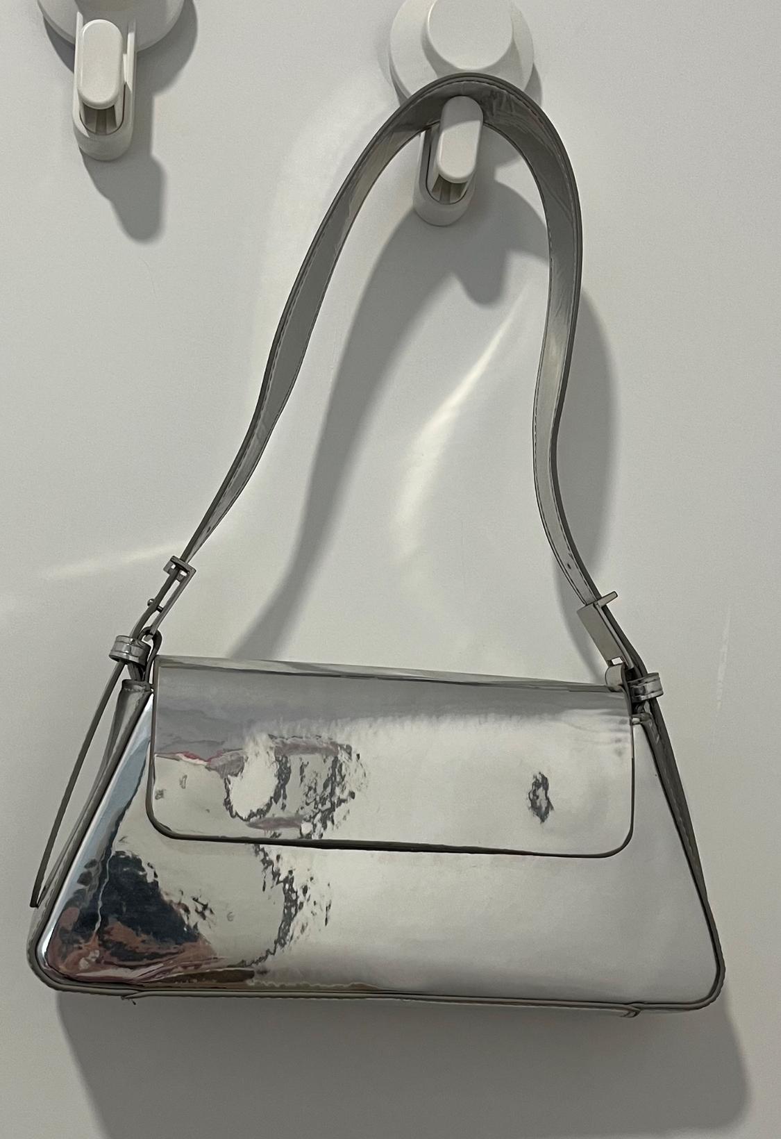 Silver metallic shoulder bag hanging on a white hook on a white wall, reflecting light and shadows.