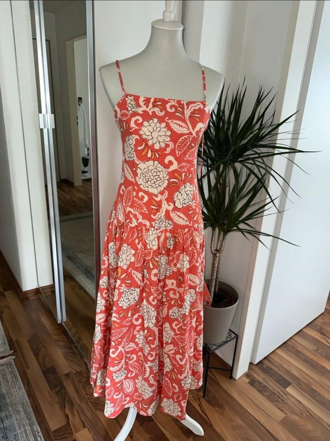 Mannequin wearing a long dress with spaghetti straps, floral print in shades of red and white, indoors with wooden floor and decorative plant.