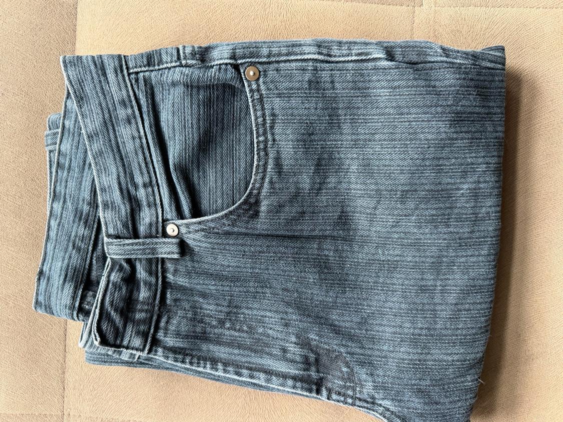 Dark grey herringbone-textured denim jeans, folded on a beige surface. Note the front pocket with a metal rivet.