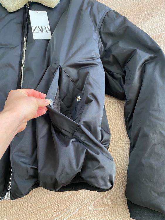 Black jacket with zipper and wool collar on a wooden floor. A hand shows the inside of the pocket. Visible tag.
