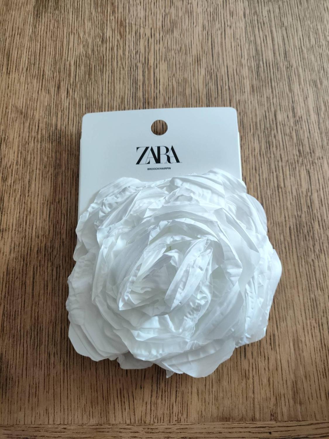 White fabric flower brooch on its label with the text ZARA BROOCH/HAIRPIN, on a wooden surface.