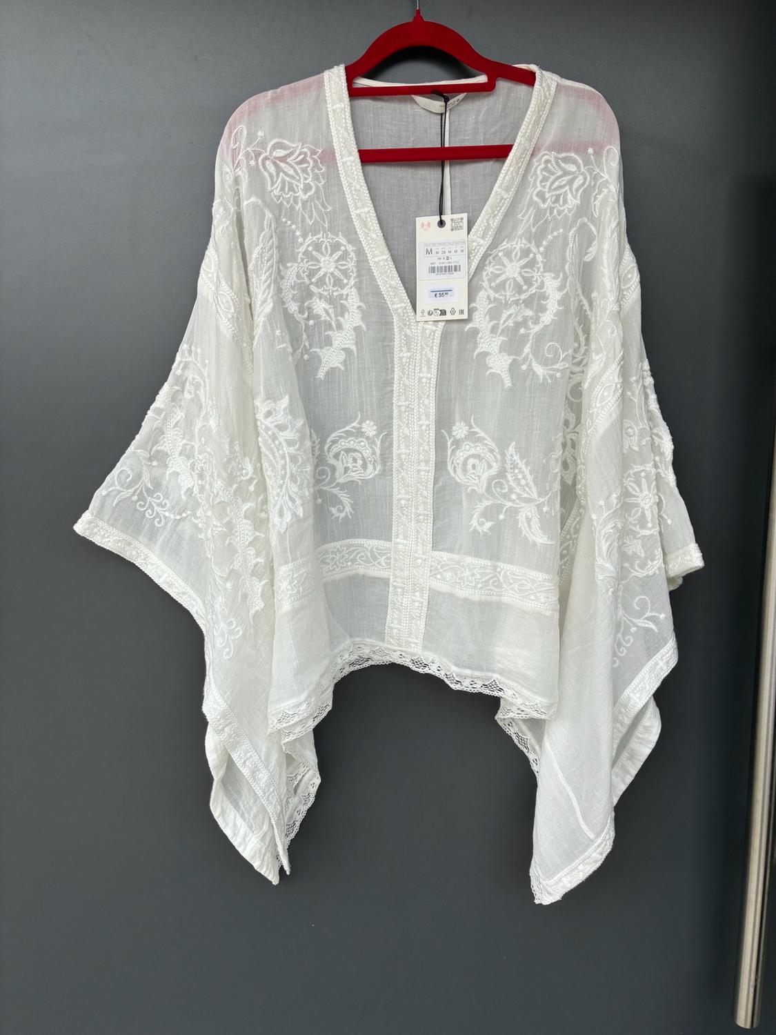 Semi-transparent white blouse with floral embroidery, voluminous sleeves and a label, hanging on a red hanger against a dark grey background.