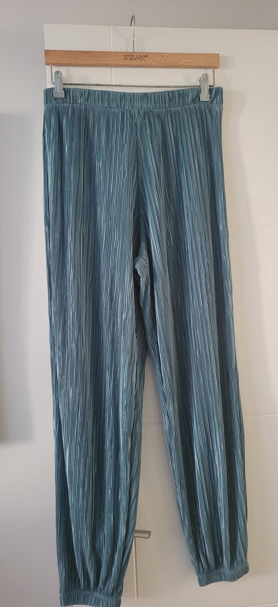 Light blue pleated trousers hanging on a wooden hanger with "United Colors of Benetton" engraved. White background.