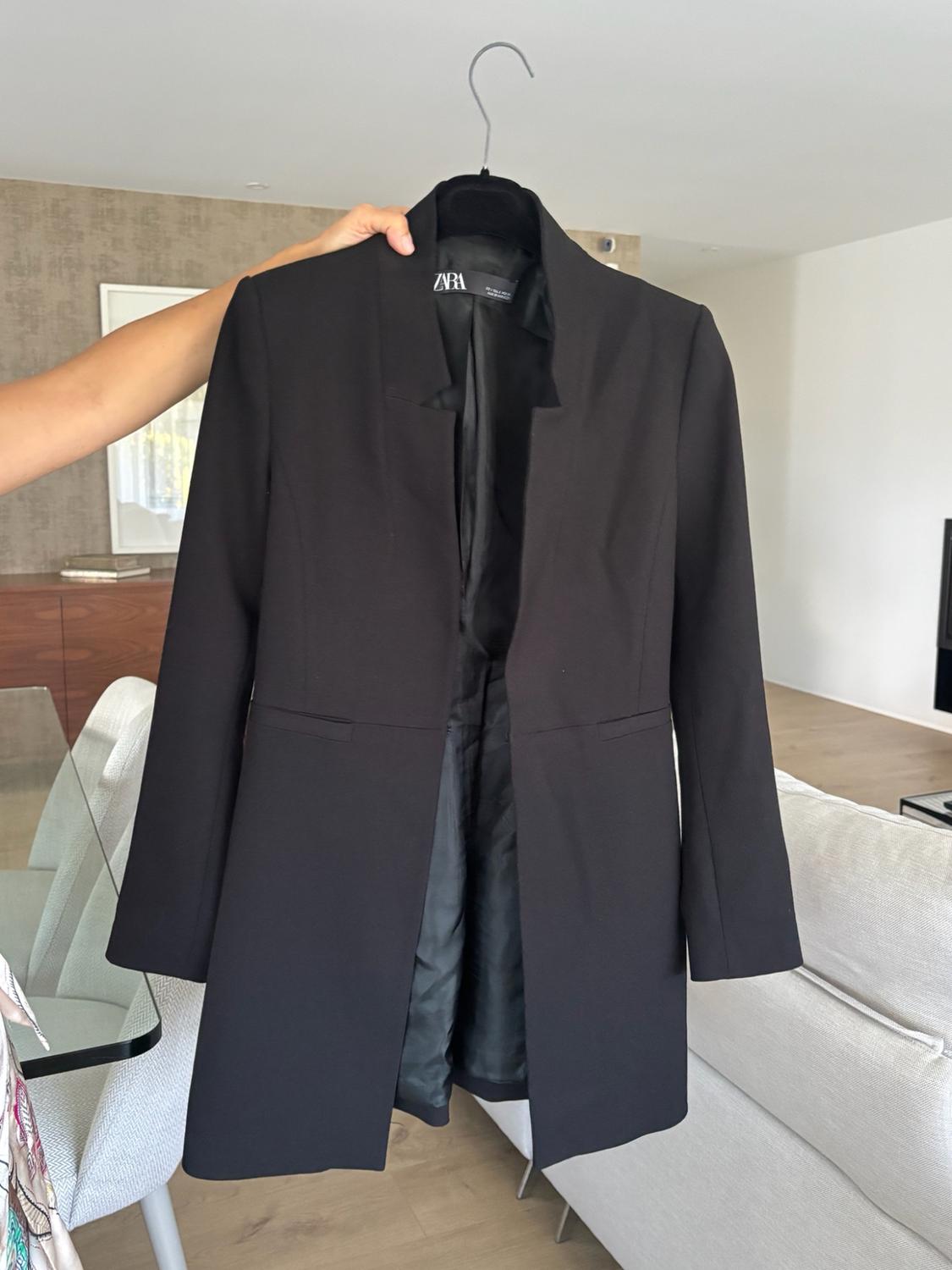 Arm holding a black jacket on a hanger with the ZARA label visible. In the background, a modern living room with a glass table and a white sofa.
