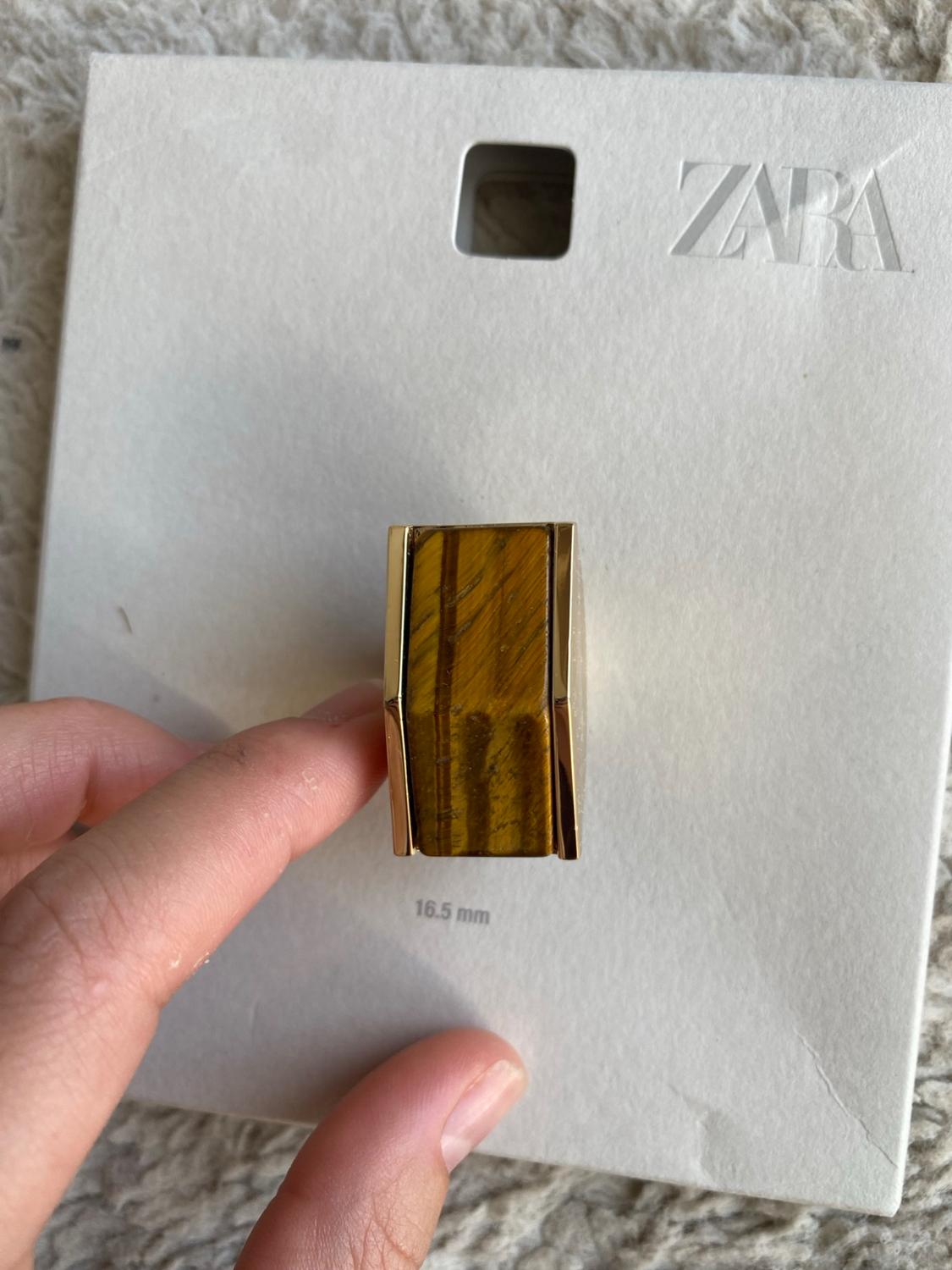 Hand holding a rectangular gold ring with a brown stone over a white label bearing the text ZARA and 16.5 mm.