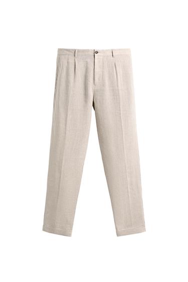 Front view of sand-coloured trousers with front pleats, side and double-welt back pockets, turn-up hems.