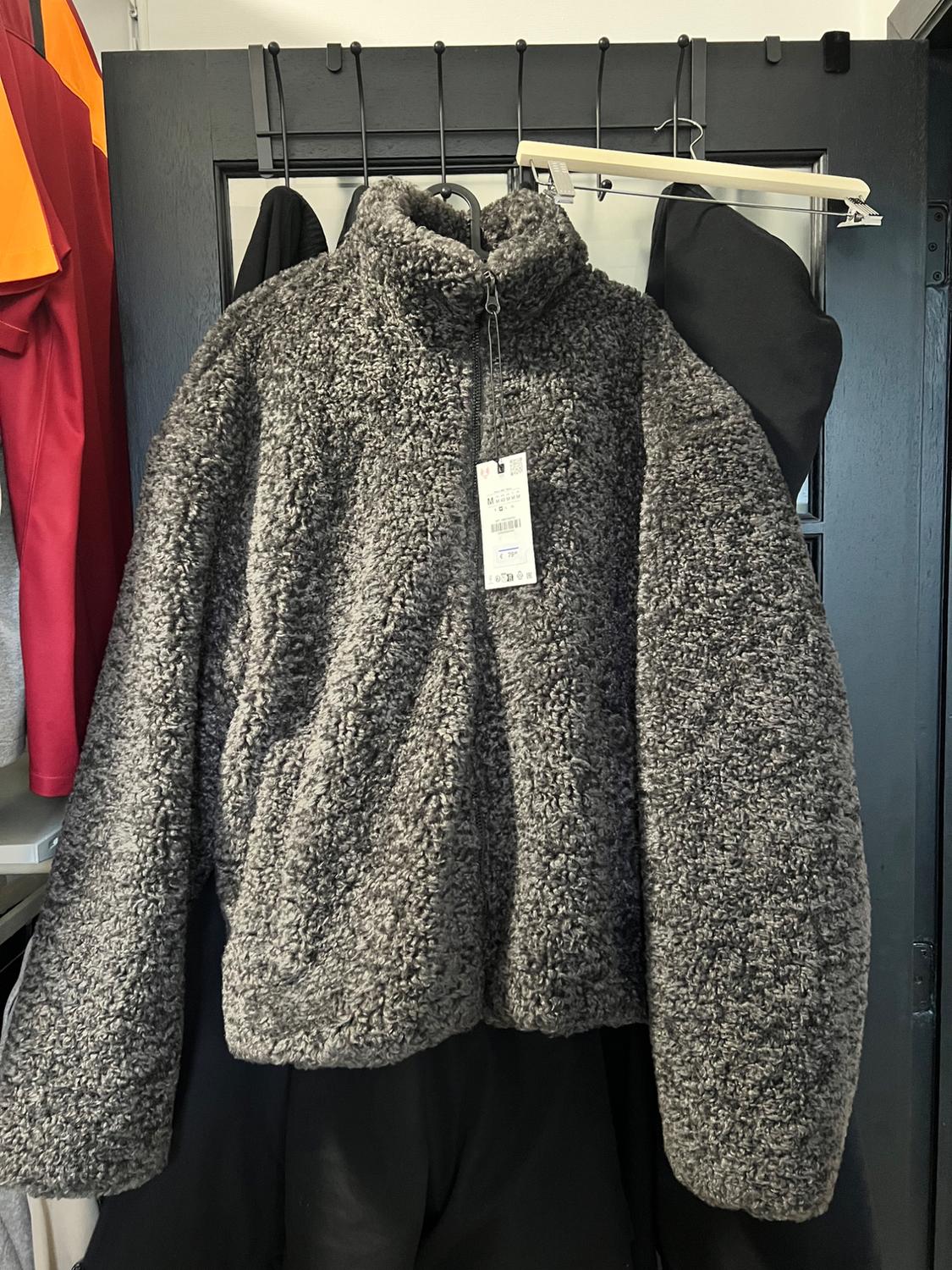 Fluffy grey jacket hanging on a black coat hanger. Visible label on the front. Background with other garments.