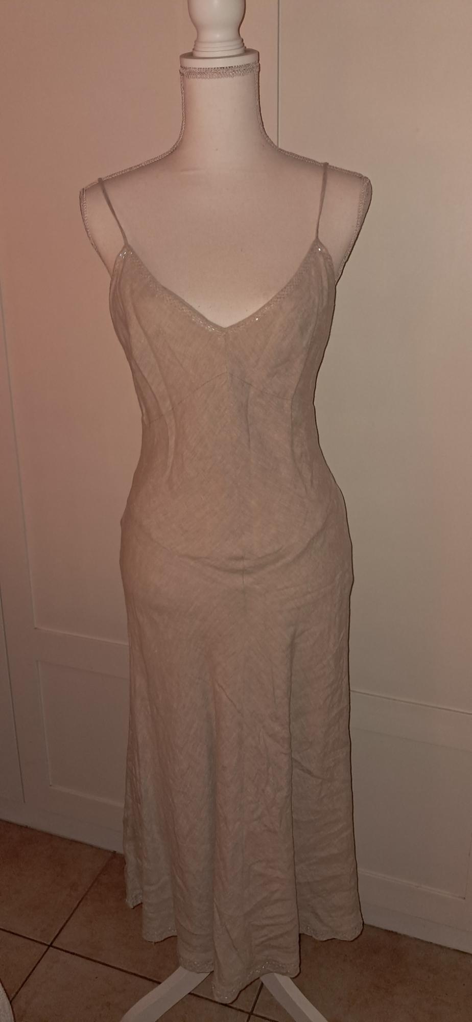 Mannequin wearing a long beige dress with thin straps and a V-neck. Relaxed indoor setting in front of a white door and tiled floor.