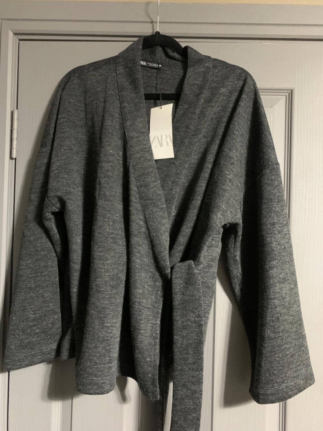 Grey long-sleeved cardigan hanging on a black hanger over a door. Visible label with text "ZARA".
