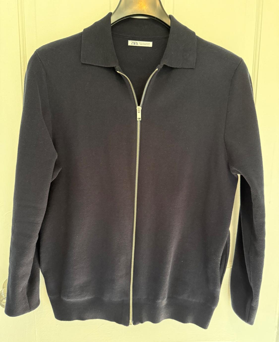 Black jacket with zip and classic collar, hanging on a hanger. Visible label on the inside of the collar. Light background.