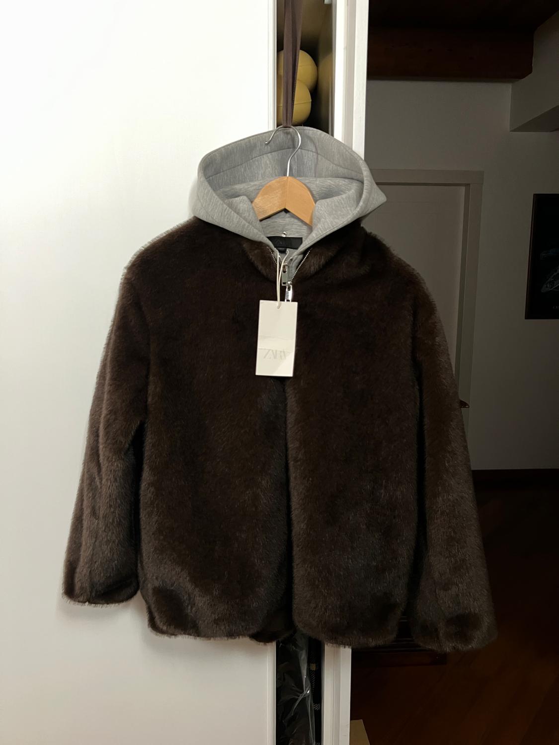 Brown faux leather jacket with a grey hood on a wooden hanger, with a visible label hanging in a white wardrobe.