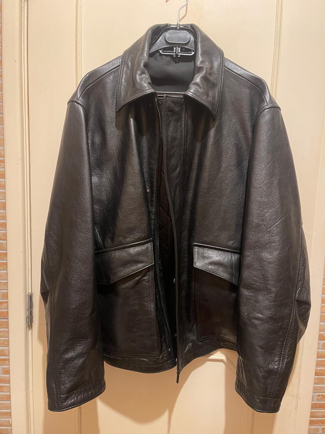 Black leather jacket hanging on a hanger over a beige door. The jacket has large pockets and a front fastening.