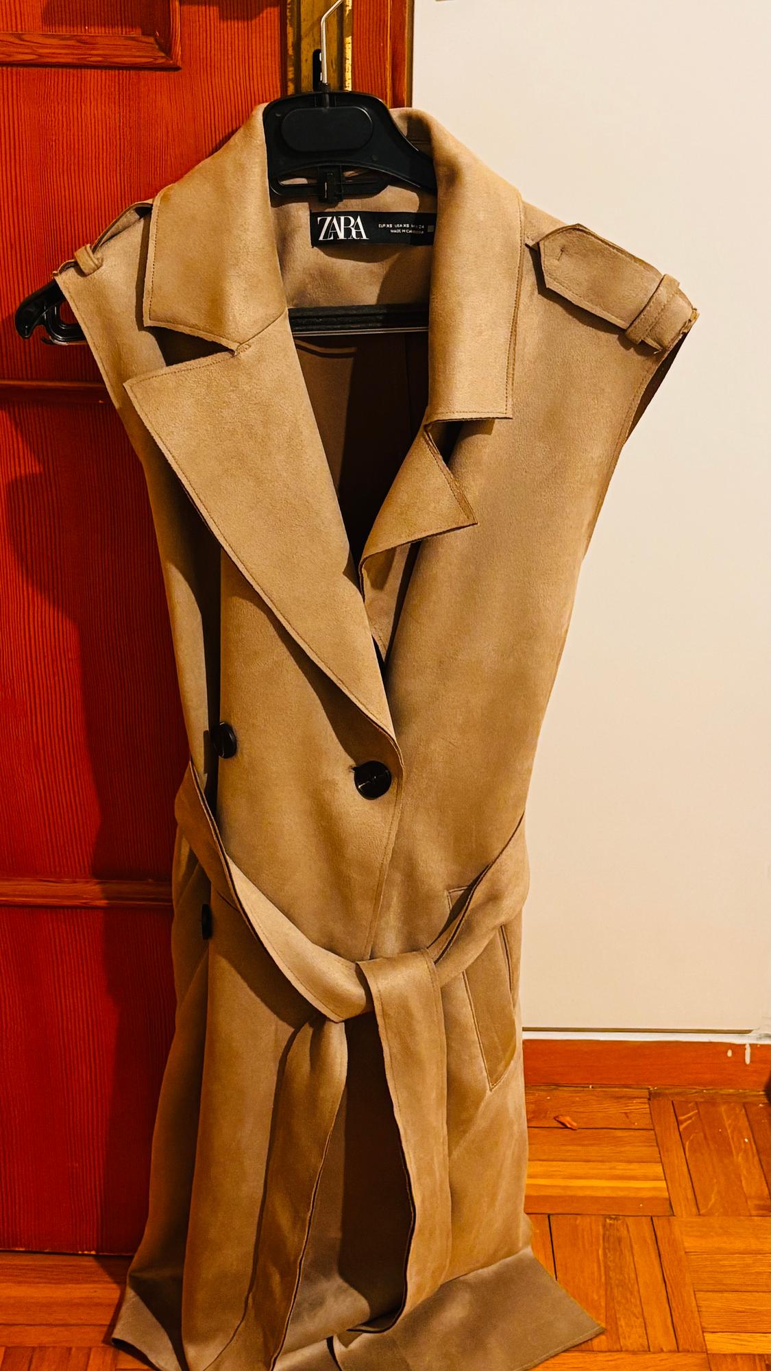 Beige sleeveless coat hanging on a black hanger, with a black belt and buttons, in front of a red wooden door. "ZARA" label.