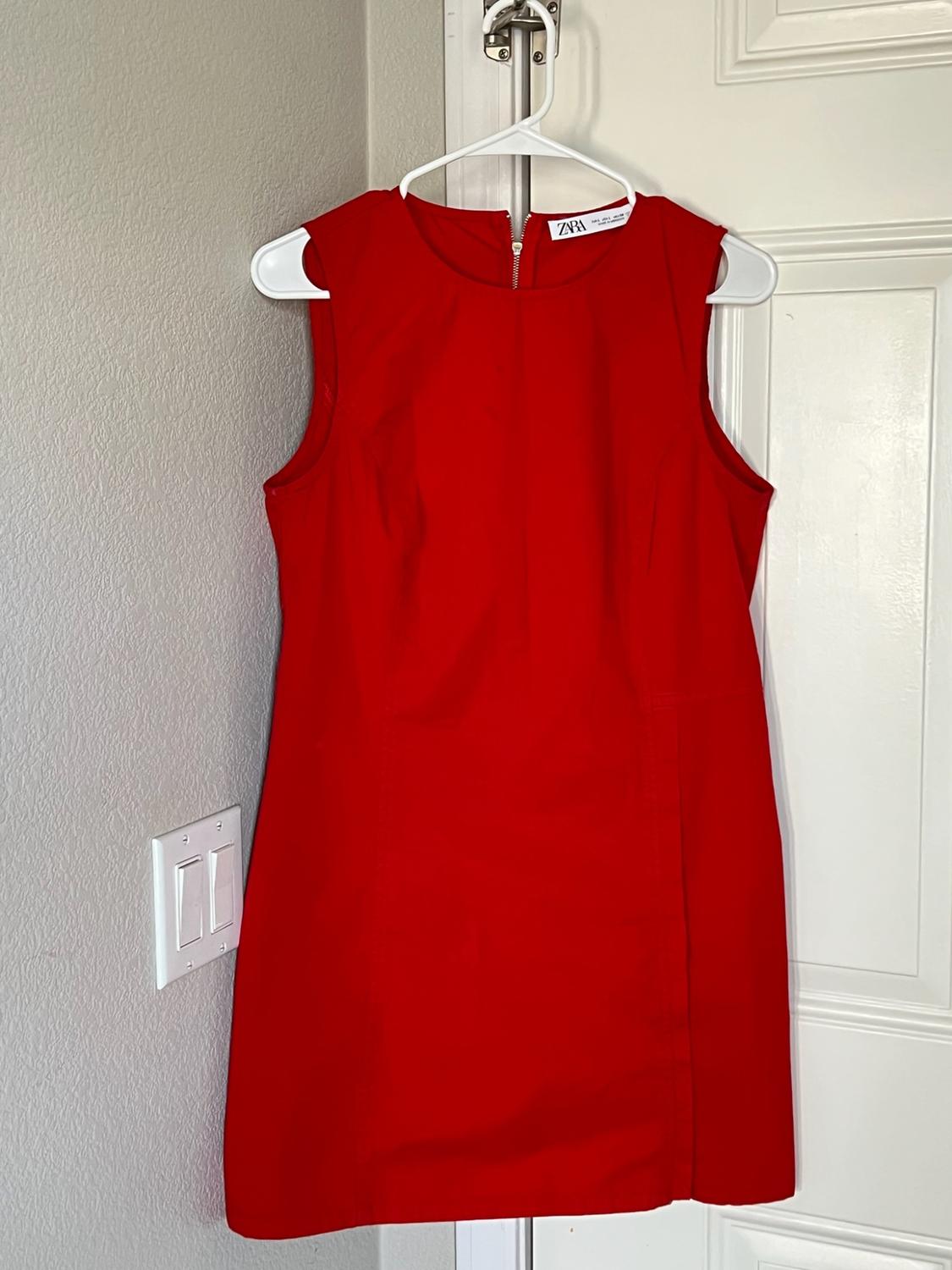 Red sleeveless dress hanging on a white door. Tag with ZARA text. Casual home setting with a gray wall and light switch.