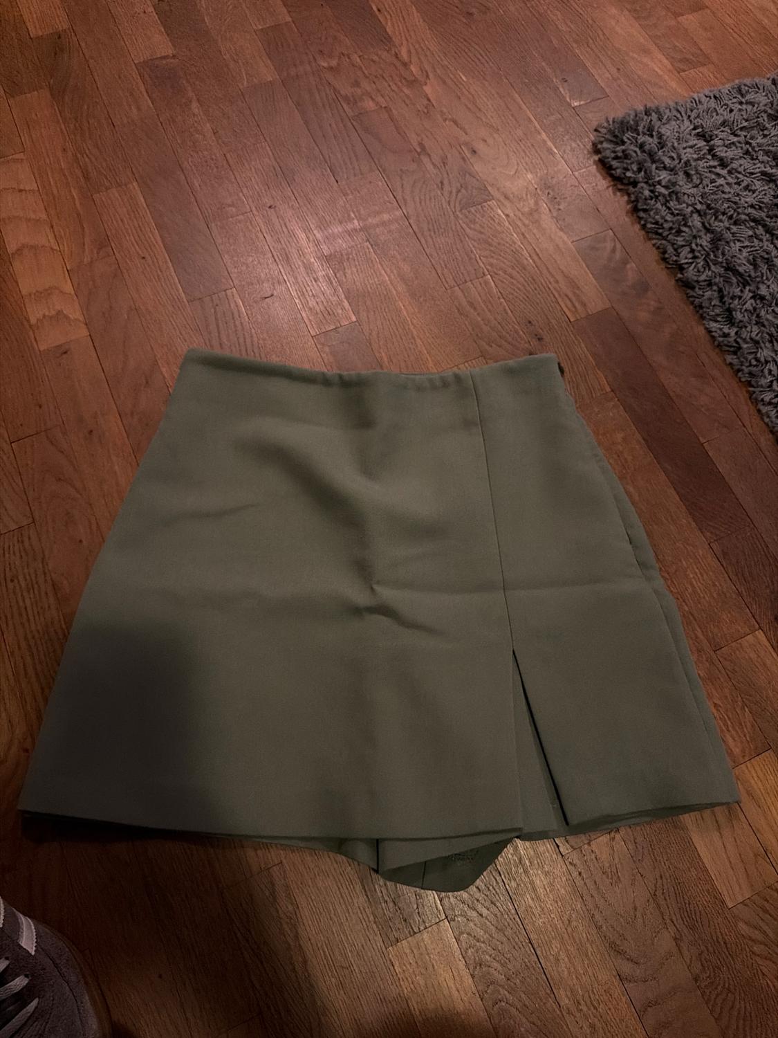 Olive-green skort with a split, on a wooden floor. Part of a grey rug can be seen.