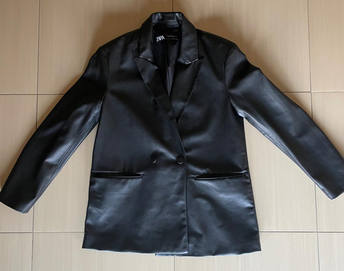 Black faux leather jacket with lapels and two buttons, laid out on a beige tiled floor. Label visible inside.