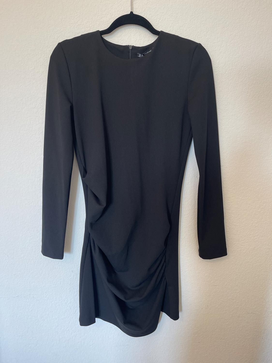 A black long-sleeved dress with draping at the front, hanging on a black hanger against a white wall.