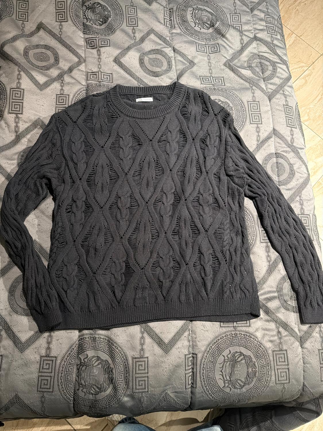 Black knitted jumper with argyle design and openwork texture, on a grey bedspread with geometric and medallion patterns.