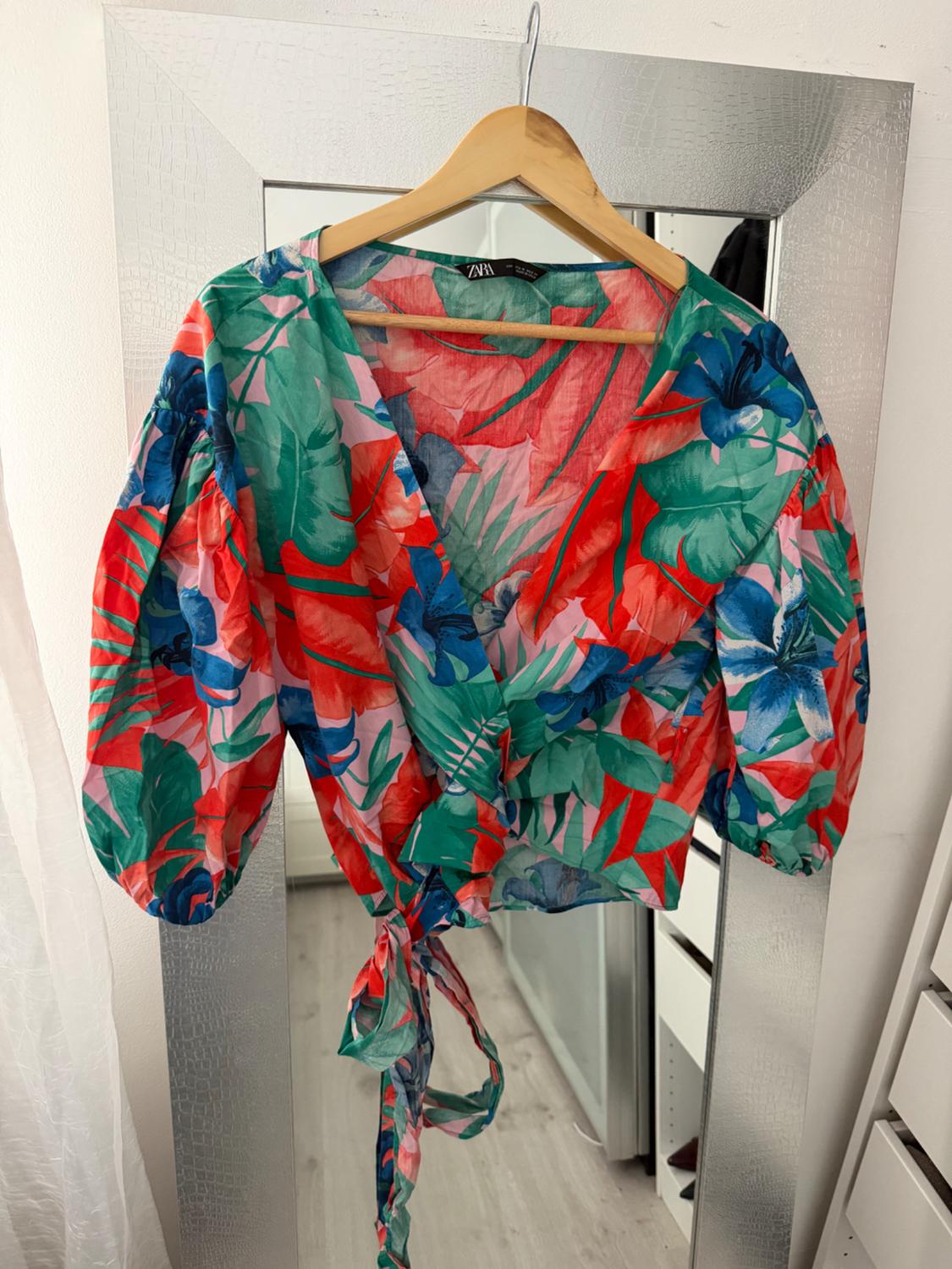 Colourful blouse with a floral print in red, green, and blue tones, hanging on a hanger in front of a mirror.