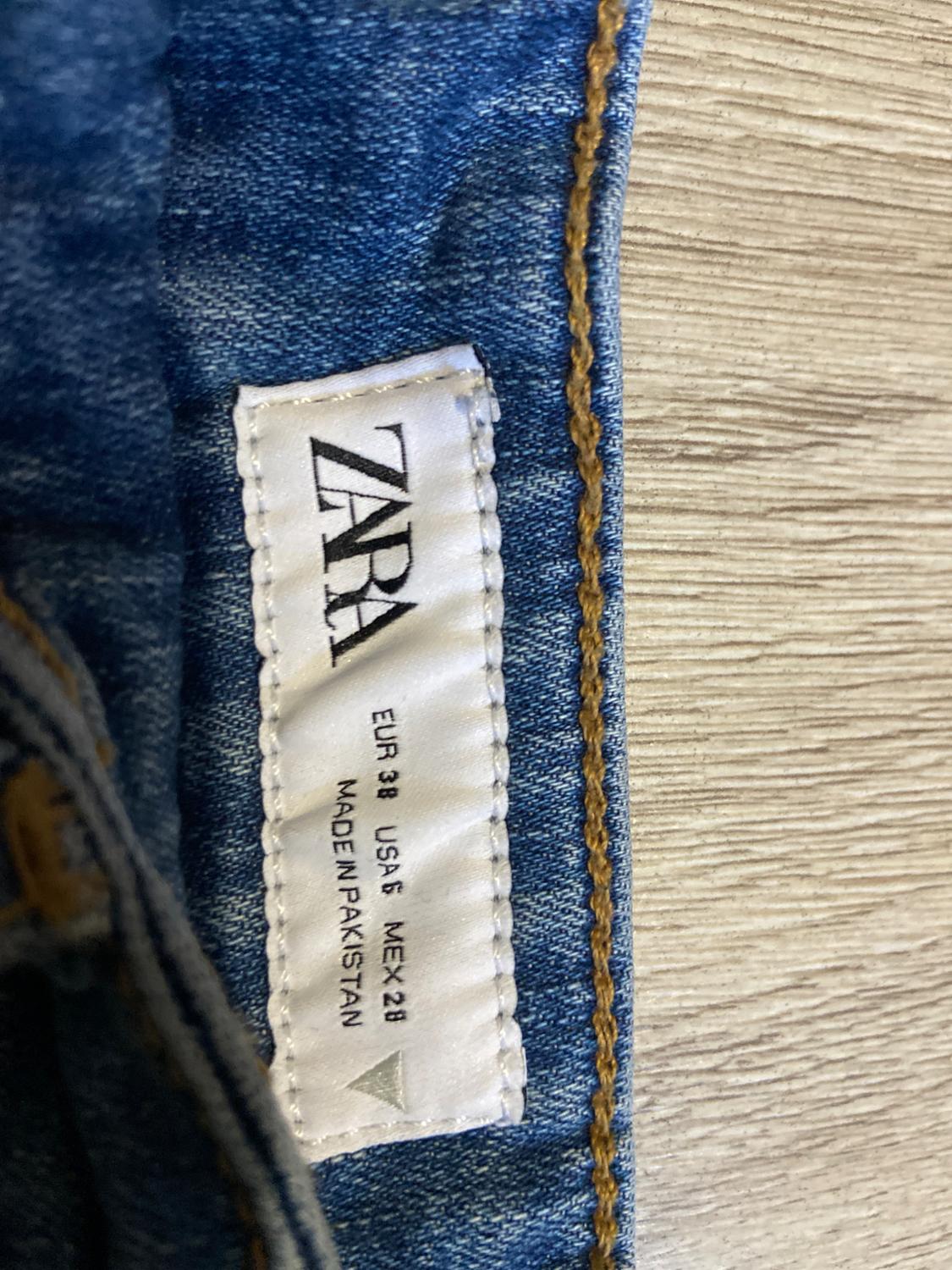 White fabric label on blue jeans with text: "ZARA, EUR 38, USA 6, MEX 28, MADE IN PAKISTAN". Light wood background.